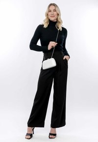 Suéter negro de punto acanalado con cuello de tortuga, pantalones de pierna ancha negros y tacones abiertos negros, sosteniendo un pequeño bolso blanco brillante con una cadena.