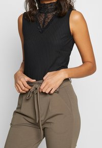 Bodysuit sans manches noir à côtes avec un col en dentelle haut et un devant en V. Associé à un pantalon marron clair avec taille tied.