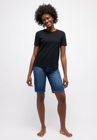 Zwarte katoenen t-shirt met korte mouwen, gecombineerd met knielange blauwe denim shorts. Model is op blote voeten en poseert op een witte achtergrond.