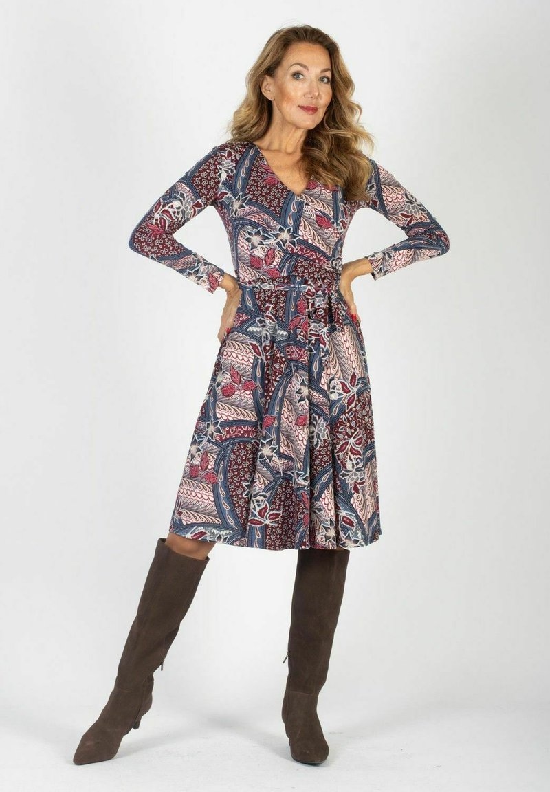 Kobieta z długimi falującymi włosami, ubrana w wzorzystą sukienkę do kolan i brązowe wysokie botki, stojąca z rękami na biodrach na białym tle.