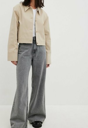 Kobieta ubrana w beżową krótka kurtkę na białym topie, w wysokim stanie szerokie szare dżinsy i czarne masywne buty, stojąca na jednolitym tle.