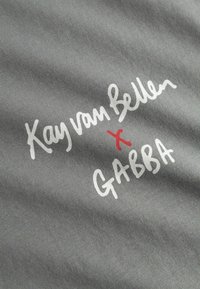 Šedá tkanina s bielym a červeným ručne písaným textom: "Kay van Bellen x GABBA." Mäkká textura, ležérny štýl, bez ďalších vzorov alebo kovania.