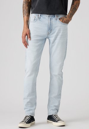 Homme portant un jean bleu clair, un T-shirt noir et des baskets basses noires avec des lacets blancs, debout avec une main dans la poche.