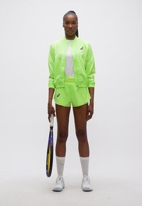 Γυναίκα με αθλητικό μπουφάν σε νεον πράσινο και σορτς, λευκά αθλητικά παπούτσια και κάλτσες, κρατώντας ρακέτα τένις, στέκεται μπροστά σε ένα απλό φόντο.