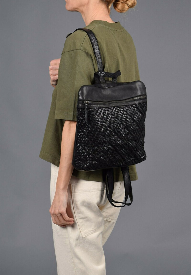 Sac à dos en cuir tressé noir avec un haut zippé, une silhouette plate et des bretelles doubles, associé à un haut vert olive et un pantalon beige.