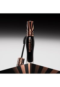 Black mascara tube with gold geometric designs and logo. Features a thick brush for volume, placed on a contrasting striped background.