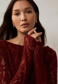 Top in pizzo color borgogna con maniche lunghe, caratterizzato da un delicato motivo floreale e un design scollato sulle spalle, abbinato a orecchini a cerchio discreti.