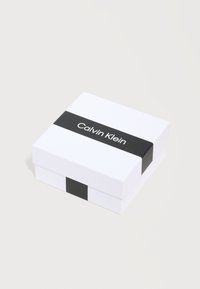Λευκό τετράγωνο κουτί με μαύρη λωρίδα που εμφανίζει το λογότυπο "Calvin Klein", τοποθετημένο σε ανοιχτό ουδέτερο φόντο.