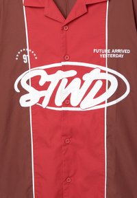 Lyhythihainen paita punaisesta ja ruskeasta kankaasta, jossa on suuri valkoinen "STWD" -logo ja teksti "TULEVAISUUS SAAPUI EILEN" valkoisella brodeerauksella.