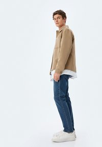 Chaqueta beige con cuello y bolsillo frontal, superpuesta sobre una camisa blanca, combinada con jeans azules y zapatillas blancas, de pie contra un fondo liso.