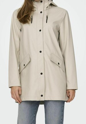 Femme portant un imperméable à capuche beige avec des boutons-pression noirs et des poches à rabat, assorti à un jean bleu clair.