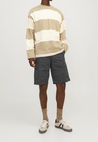 Uomo che indossa una camicia a maniche lunghe beige e bianca a righe, pantaloni cargo grigio scuro, calze beige e sneaker beige su uno sfondo neutro.