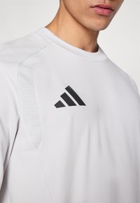 Λευκό αθλητικό μπλουζάκι με στρογγυλή λαιμόκοψη, απαλό ύφασμα, διατρητικές λεπτομέρειες στους ώμους και μαύρο λογότυπο adidas στο στήθος.