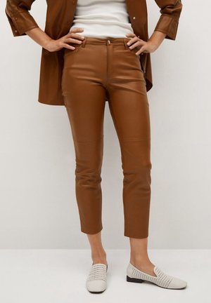 Person wearing cropped brown leather pants, white woven slip-on shoes, white top, and brown button-up jacket with hands on hips.