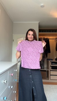 Vijolična majica s kratkimi rokavi z majhnim belim vzorcem, ki ima ohlapno prileganje. Pod njo je oblečena temno siva krila z gumbi.