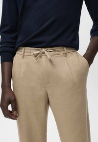 Pantalons en lin beige avec une taille à cordon, poches latérales et une coupe décontractée, associés à un pull bleu marine. Texture lisse, tissu léger.