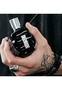 Bouteille de parfum en forme de poing noir avec un capuchon argenté et le logo « DIESEL » en relief, tenue dans une main tatouée, arborant une bague et un bracelet en argent.