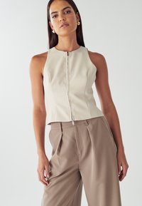 Top senza maniche, beige, con zip e design aderente, abbinato a pantaloni beige a vita alta, plissettati. Tessuto morbido e stile minimalista.
