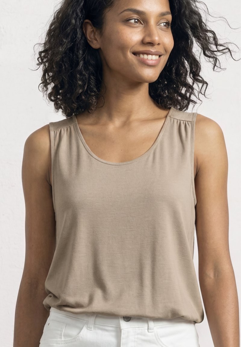 Femme souriante aux cheveux bouclés portant un haut beige sans manches et un pantalon blanc, regardant sur le côté sur un fond uni.