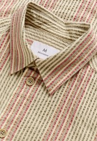 Camisa de botones en beige con rayas alternas en rojo y marrón. Presenta una tela texturizada y un cuello estándar con cierre de botón.