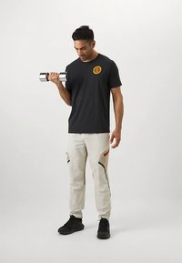 Juodas marškinėliai su geltonu emblemų, kreminės spalvos kelnės su juodais ir raudonais akcentais, laiko sidabrinį hantelį, dėvi juodus sportbačius.