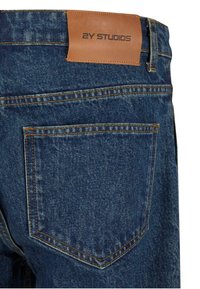 Denim jeans with a dark indigo wash, featuring a tan leather label at the waistband and contrasting yellow stitching on the back pocket.