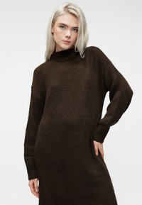 Blonde Frau in einem dunkelbraunen gestrickten langen Kleid, die mit einer Hand auf der Hüfte vor einem schlichten hellen Hintergrund posiert.