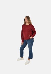 Maglione a maglia di colore cremisi con scollatura rotonda, polsini e orlo a costine, caratterizzato da un logo a forma di cuore sul davanti; abbinato a jeans blu e scarpe da ginnastica bianche.
