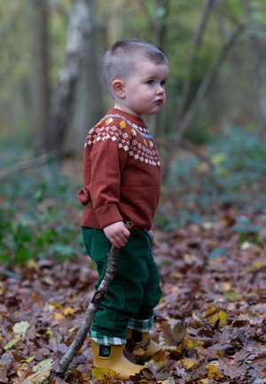 Ein Kind trägt einen rostfarbigen gestrickten Pullover mit floralen Mustern, grüne Hose und gelbe Stiefel, während es einen Stock in einer Waldumgebung hält.
