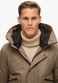 Tan struktureret frakke med en stor krave med sorte kanter, der har sidelommer og en lys beige turtleneck sweater under.