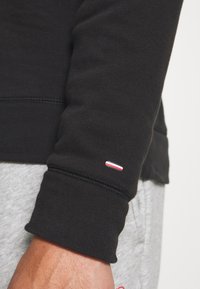 Sudadera negra de tela con puños acanalados. Pequeño logo en rojo, blanco y azul en el puño. Textura suave y diseño ajustado.