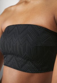 Juodas bandeau tipo viršus su tekstūruotu zigzagu raštu, pagamintas iš elastinės medžiagos. Be dirželių; yra tvirtinimo elastinė juosta. Detali artima dizaino apžvalga.