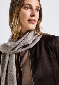 Ljust grå, mjuk tygscarf hängande runt halsen, i kombination med en mörkbrun bomberjacka och en beige topp med en slät textur.