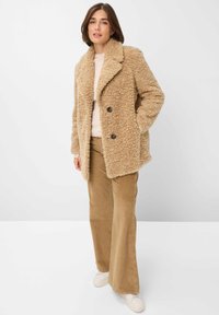 Beige neplederen bontjas met een notch-kraag, dubbelgeknoopte knopen en zijzakken. Gekleed met een lichtbeige trui en corduroy broeken.