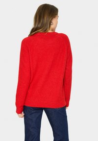 Roter Pullover mit einer weichen Stricktextur, Rundhalsausschnitt und geripptem Saum. Über dunkelblauen Jeans getragen, von hinten betrachtet.