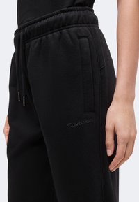 Κοντινό πλάνο μαύρων παντελονιών φόρμας Calvin Klein με ρυθμιζόμενο κορδόνι στη μέση και πλαϊνές τσέπες, που φοριούνται από άτομο με ένα εμφανές χέρι.