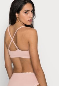 Bralette rosa claro com design cruzado nas costas, feito de tecido suave. Características notáveis incluem alças ajustáveis e uma faixa elástica suave.