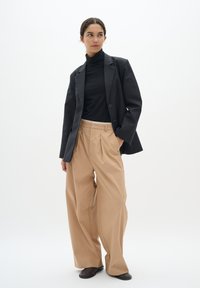 Musta bleiseri mustan turtleneckin päällä, yhdistettynä beigeihin leveälahkeisiin housuihin. Sileä kangas, hyvin istuva leikkaus, joissa on hienovaraiset laskokset ja minimaalista metallisosaa.