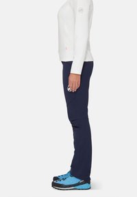 Seitenansicht einer Person, die ein weißes Langarmshirt, marineblaue Hose und blaue Wanderschuhe trägt, mit Logoschildern auf dem Arm und der Hose.