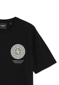 T-shirt en coton noir avec un logo rond aux accents dorés, texte "Né aux Frontières", et coupe décontractée. Manches courtes, design col rond.