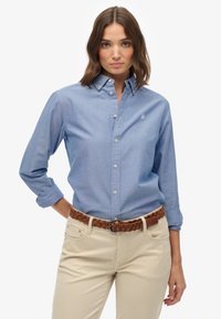 Jasnoniebieska koszula z długim rękawem, z klasycznym kołnierzykiem i małym haftowanym logo, w połączeniu z beżowymi spodniami i brązowym plecionym paskiem.