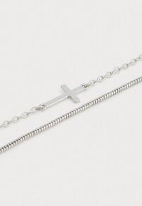 Δύο ασημένιες αλυσίδες σε λευκή επιφάνεια, η μία με ένα οριζόντιο σταυρό και η άλλη με λεπτή αλυσίδα φίδι.