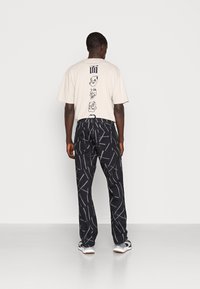 Beige t-shirt with graphic print, black pants with white geometric pattern. Casual fit, cotton material, paired with gray and white sneakers.