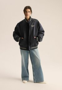 Granatowa kurtka bomber z białymi paskami na mankietach i dolnym zakończeniu, z haftowanym napisem. W zestawie z jasnoniebieskimi, szerokimi dżinsami.