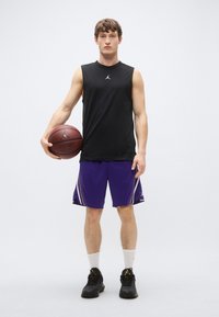 Tricou negru fără mâneci cu un logo, pantaloni scurți mov cu dungi galbene, ținând un baschet, încălțat cu pantofi negri și șosete albe.