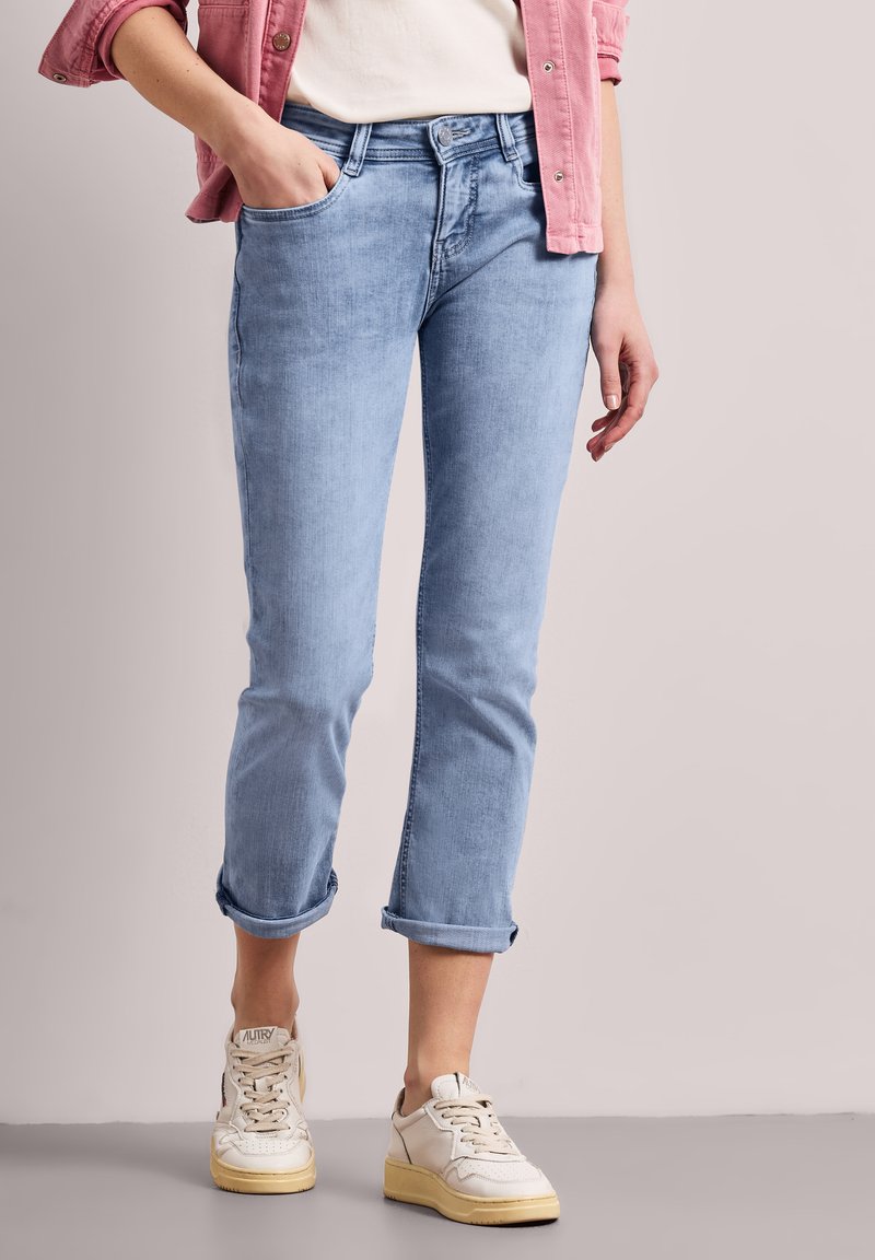 Femme portant un jean bleu clair retroussé, des baskets blanches, une chemise blanche et une veste rose avec une main dans la poche.
