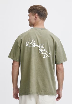 Oliven grøn bomuld t-shirt med en hvid stregtegning af en hånd, der hælder fra en flaske i et glas på ryggen.