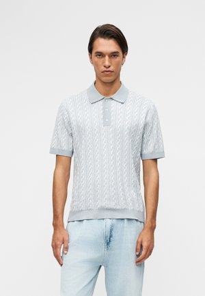 Jonge man die een lichtblauw patroon polo met korte mouwen en lichtgewassen spijkerbroek draagt, staand tegen een effen achtergrond.