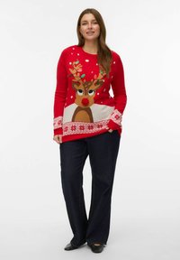 En kvinna som står iförd en röd jultröja med ett ren- och snöflinge-mönster, mörka vida jeans och svarta skor, leende.