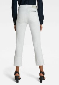Jeans de mezclilla blanca acampanados con una cintura alta, que cuentan con bolsillos traseros, un bolsillo con cremallera y puños doblados. Combinados con sandalias de tacón negras.